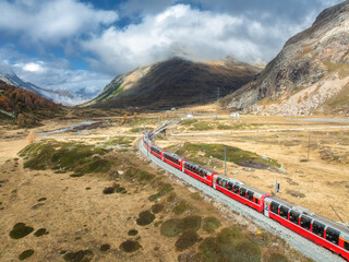Wall Mural - aerial view of modern red train, alpine mountains, orange hills and trees, sky with clouds at sunny 