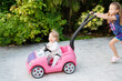 © Ana - Happy baby in a pink toy car smiles while gripping the steering wheel, as an older sister joyfully pushes from behind capturing sibling fun and outdoor playtime. Authentic, candid shots