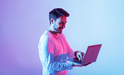 A young man stands with a laptop, engaged in his work. The background is illuminated with vibrant pink and blue hues, creating a dynamic and modern atmosphere.
