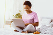 © Prostock-studio - Remote work and viewing webinar from home. Smiling african american girl sitting on bed with laptop, croissant on plate near, sun flare, free space