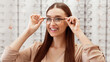 © Prostock-studio - A young woman is smiling while adjusting her eyeglasses in an optical store filled with various frames. She appears excited about her new look and options available.