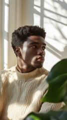 Aufkleber - Young man in a cream sweater gazes thoughtfully by a sunlit window with plants in a modern indoor setting