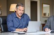© SimpleDesignStudio - Middle-aged Caucasian man focused on work at desk with laptop, papers, and serious expression in cozy office setting