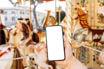 Mockup woman's hand holding using smart phone with white blank screen POV on the background of a carousel with horses, mobile app design and advertising, online marketing