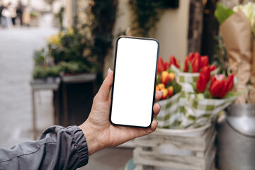 mockup woman's hand holding using smart phone with white blank screen pov at flower street shop, mob
