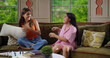 © Raushan_films - Indian happy gen z young adult two woman lady sitting cozy sofa couch enjoy day indoor house teen age female cute girl child hold pillows have funny fight battle hit each other fun joy spend time