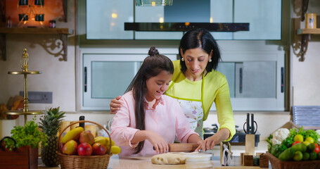 Indian happy woman lady with cute teen age cute girl rolling raw flour dough learn cooking food indoor home smiling young mom teach to kid child baking cookies enjoy day indoor modern home kitchen