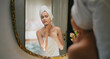 © Raushan_films - Indian young adult gen z woman lady wrapped towel on head and body look mirror apply fresh make up cream on dry face teen age girl female do gentle facial massage day skin care house home