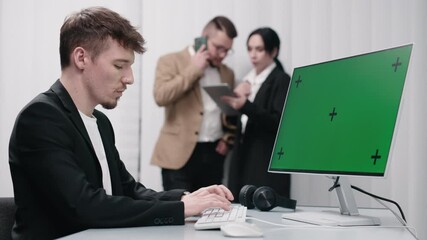  IT technician working at a desktop computer with a Chroma Key green screen, while colleagues brainstorm in the office. Concept e-commerce development, marketing, and data analysis in the office.