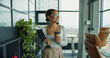 © Kostiantyn - young professional woman with glasses and coffee cup walking confidently in a modern office, holding a laptop and smiling while looking out large glass windows with natural lighting