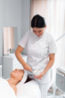 © Andrii - Dermatologist in uniform preparing face mask for female client lying on massage table at salon. Relaxed young woman undergoing facial treatment in beauty spa