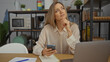 © Krakenimages.com - Woman working in office holding phone and thinking at desk with laptop, notebook and kitchen background, showcasing modern workplace and thoughtful expression.