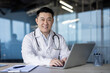 © Tetiana - Portrait of a smiling young Asian male doctor professional working in a hospital, sitting in the office at a table with a laptop and looking at the camera