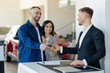 © Anastasiya - A cheerful young couple receives the keys to their new car from a professional salesman at modern dealership. Man smiling to salesperson, and the woman stands happily beside him