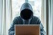 ©  Studio AB Images - Front view of internet scammer sitting in hood with hidden face using laptop on the table in room in daytime