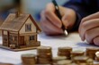 © shirina design - A person is writing on a document while a model house sits next to stacks of coins.