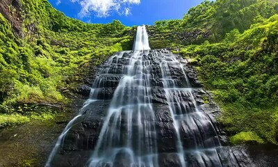 Wall Mural - Majestic Waterfall Cascading Down Lush Green Cliffs Under a Bright Blue Sky with Fluffy White Clouds