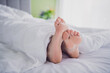 © deagreez - Close-up of two feet peeking out of a cozy white duvet on a bright airy morning, creating a mood of relaxation and comfort.