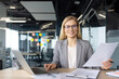© Liubomir - A businesswoman is working at her desk with a laptop and holding documents, smiling at the camera in her office