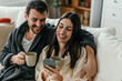 © La Famiglia - Happy couple laughing and using smartphone while drinking coffee on sofa