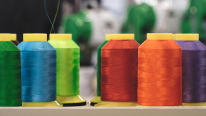 Aufkleber - A row of colorful spools of thread are displayed on a shelf. The spools are of various colors, including green, yellow, orange, and purple. The spools are arranged in a neat row