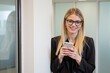 © Nina - Confident Young Caucasian Woman with Long Blonde Hair and Glasses Checking Her Smartphone in a Modern Office Environment