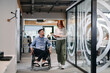 © .shock - Business colleagues, collaborative business colleagues, including a person in a wheelchair, walk past a modern glass office corridor, illustrating diversity, teamwork and empowerment in the workplace.
