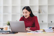 © kenchiro168 - Excited Asian businesswoman in red sweater working on laptop at desk surrounded by documents and coffee cup.