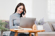 © PRIME STOCK LAB - Asian businesswoman smiling while talking on phone and working on laptop in cozy home office setting.
