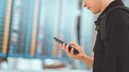 Close-up of a man passenger using phone at airport terminal, Checking boarding time online on smartphone, flight information screens in background, travel concept.