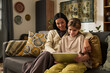© pressmaster - Mother and daughter smiling while using tablet together on a cozy living room sofa. Decorated in warm tones with patterned cushions and a tapestry wall hanging