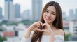 © rdkcho - Charming Young Asian Woman with Long Brown Hair Makes Heart Shape with Hands, Cityscape Background, Romantic and Sweet Expression, Close-Up Portrait