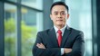 © STK - Portrait of a confident Asian businessman in a dark suit and red tie, arms crossed, standing in a modern office with blurred background.
