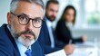 © STK - Portrait of a middle aged man with salt and pepper beard and glasses, in a business meeting. He is in the foreground with two colleagues out of focus in the background.