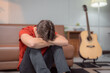 © Tj - Depressed musician is sitting on the floor and hugging his knees, suffering from mental health problems and feeling desperate and lonely in his living room
