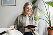 © shurkin_son - Home portrait of senior smart intelligent female in causal shirt and glasses spending morning at home, reading and drinking herbal tea on breakfast, holding book in hand, sitting next to window