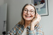 © shurkin_son - Portrait of happy laughing grandmother in glasses and plaid shirt video chatting with her grandchild online, sitting at home in living-room, having fun, enjoying nice and warm conversation