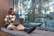 © 24K-Production - Smiling happy african american female traveler relax using laptop computer and headphones in resort with forest view.