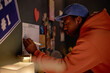 © Seventyfour - Man in orange hoodie and blue cap writing schedule on bulletin board with various items attached, in cozy room with warm light