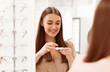 © Prostock-studio - A young woman smiles as she prepares to try on contact lenses in an optical store, surrounded by various eyeglasses on display, reflecting a moment of excitement and anticipation.