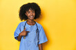 © Asier - Young African-American nurse in studio with yellow background person pointing by hand to a shirt copy space, proud and confident