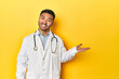 © Asier - Asian doctor in white coat with stethoscope, yellow studio showing a copy space on a palm and holding another hand on waist.