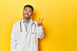 © Asier - Asian doctor in white coat with stethoscope, yellow studio joyful and carefree showing a peace symbol with fingers.