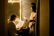 © AnnaStills - Two people in dressing room collaborating on theater script. One person seated at dressing table under warm lighting, while another person standing beside holding script pages