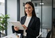 © Over View - Portrait of a smiling woman in a blazer holding a tablet in an office setting with a plant