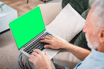 Handsome senior old man using a laptop with mock up blank screen while sitting relaxing resting on couch at home. Mature Adult man working laptop browsing internet at home copy space