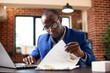 © DC Studio - Closeup of black man seated at office desk, deeply focused on analyzing financial documents in brick wall room. African american male manager examining graphs on clipboard with laptop nearby.