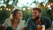 © Dulemegapixel - A happy couple sits together, laughing joyfully amid glowing lights in an outdoor setting, showcasing the warmth and connection of their relationship over drinks and conversation.