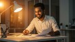 © Qbertstudio - Young adult Indian businessman smiles working at desk late at night Laptop paperwork business charts illuminate dedication and entrepreneurship