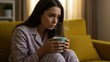© Qbertstudio - Caucasian young adult woman in pajamas holding steaming mug on couch indoors. Depression, anxiety, mental health, and self-care concepts.
